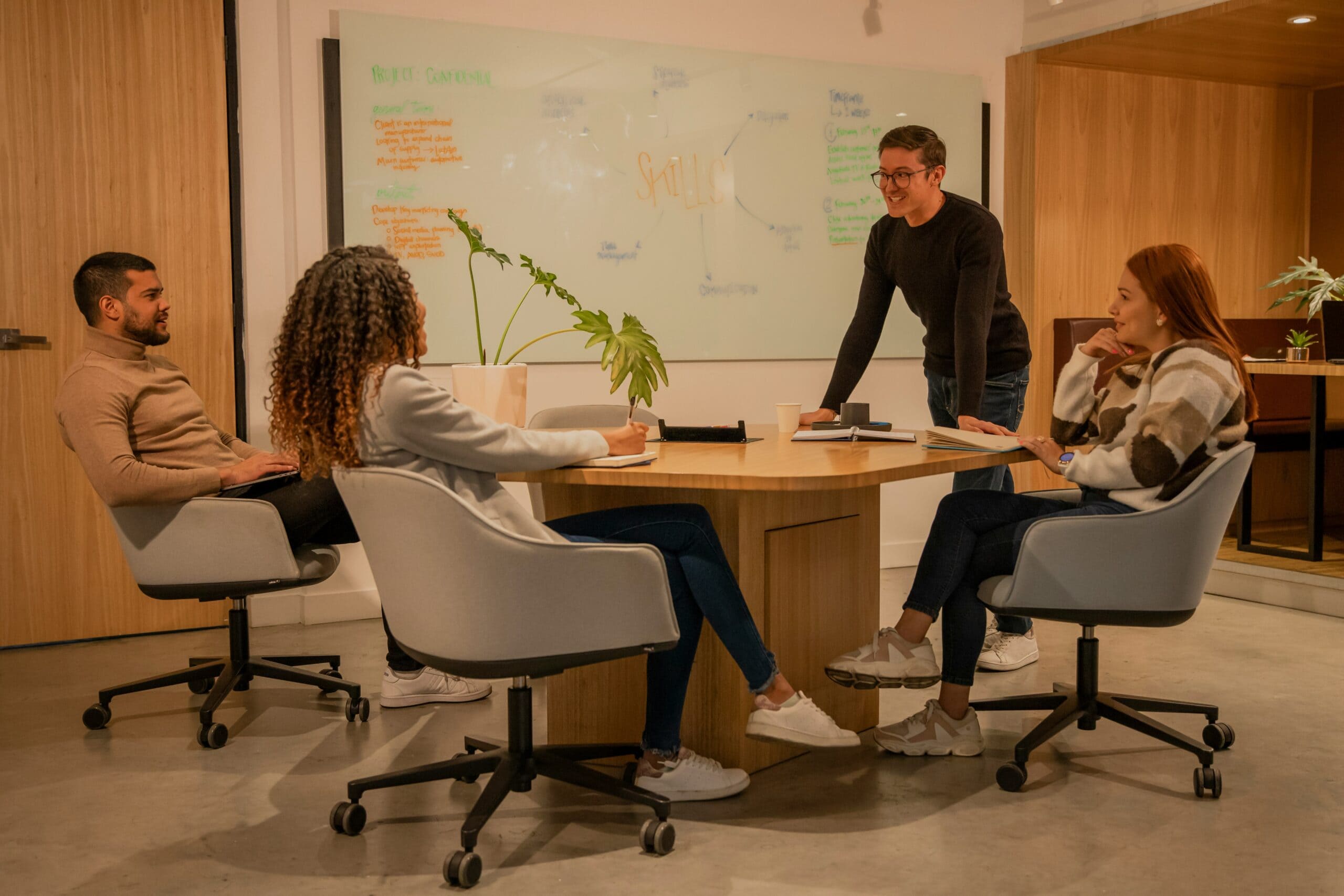 Four colleagues discussing "What makes a good IT consultant" in a modern office, one standing and pointing at a whiteboard, three others seated and engaged in the conversation.