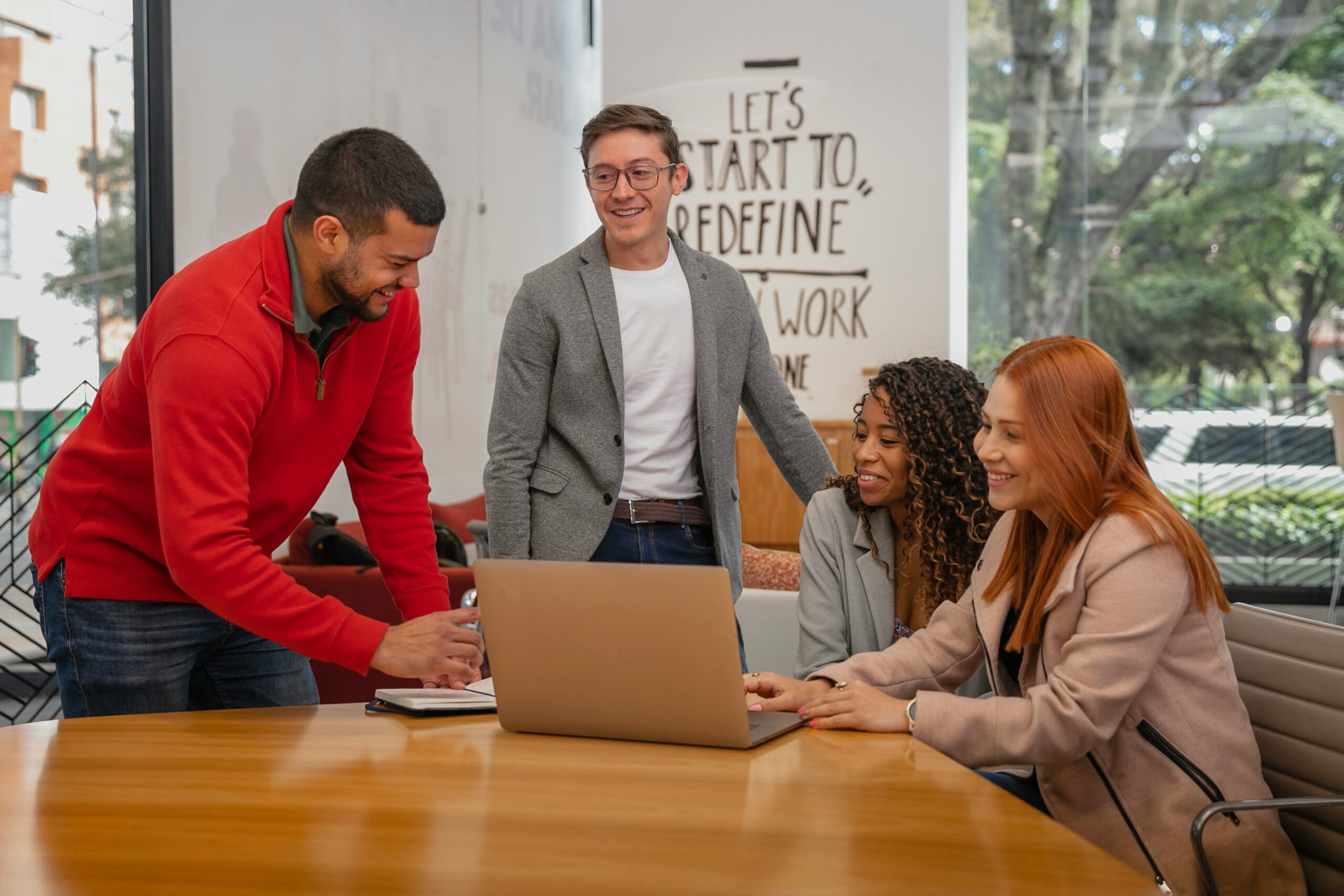 Four diverse colleagues collaborate around a laptop in a bright office, discussing what makes a good IT consultant with smiles.