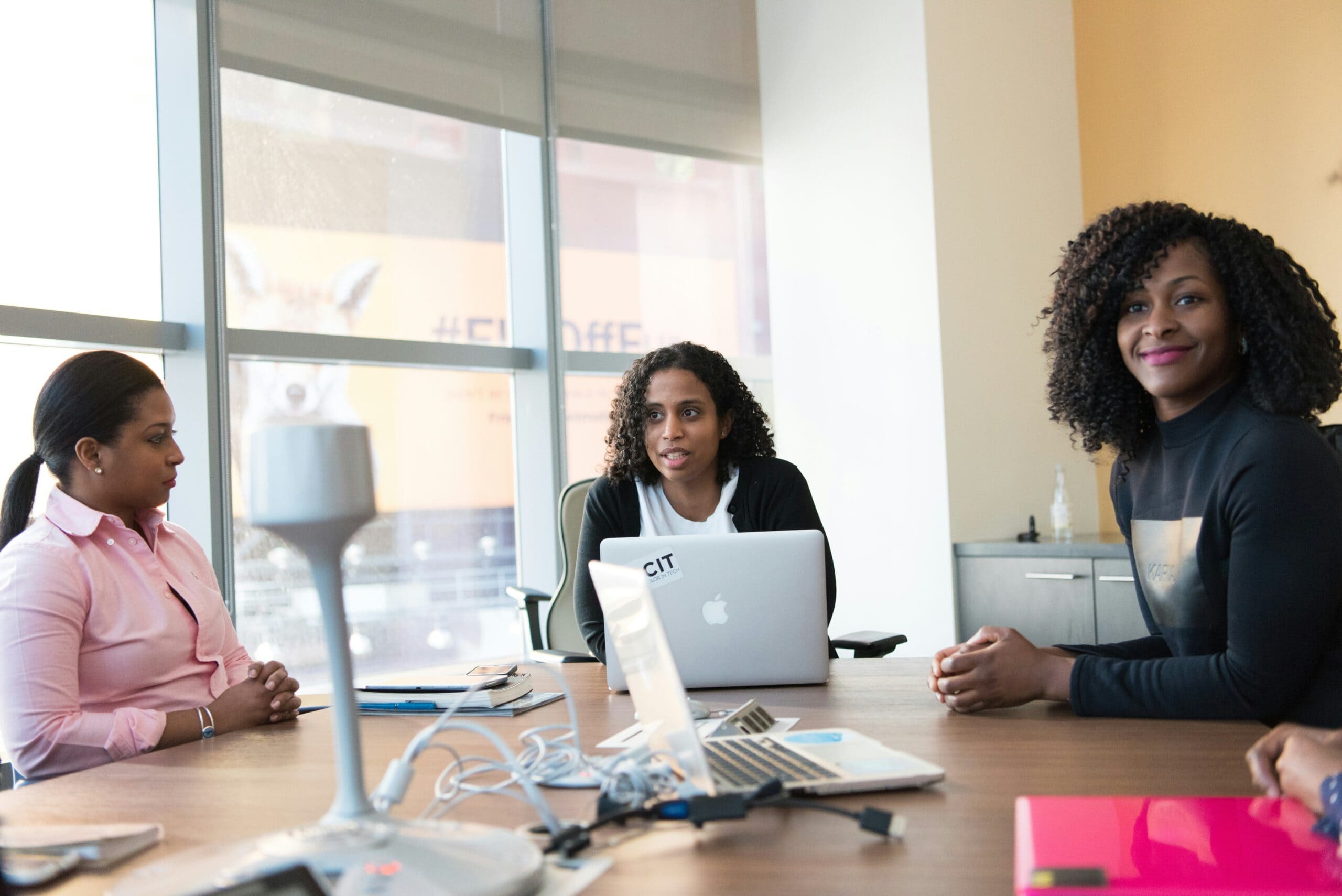 Three women seated around a conference table with laptops, engaging in a discussion about what makes a good IT consultant in an office setting.