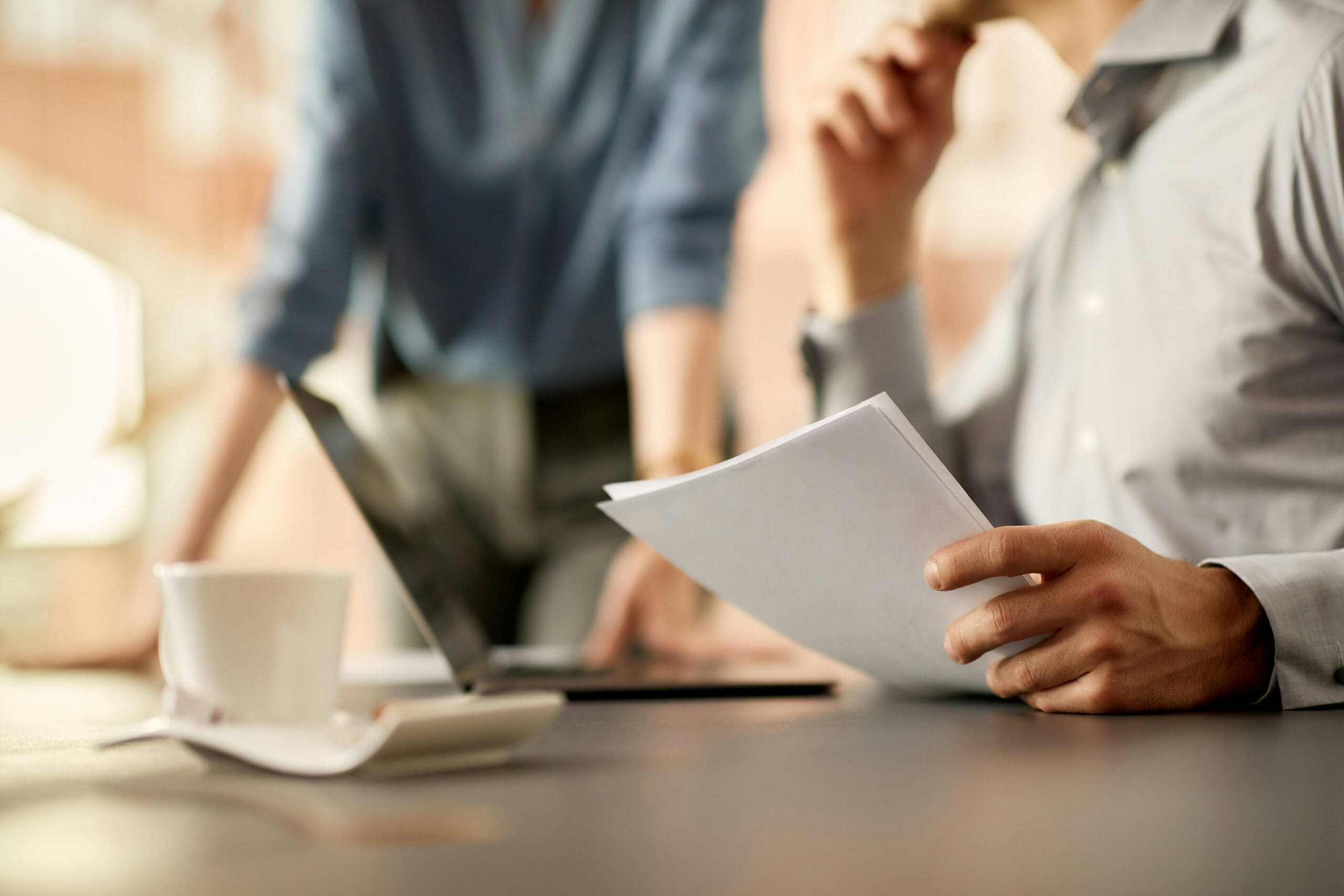 Two IT consultants discussing a document, one holding papers and the other using a laptop at a table with coffee cups.